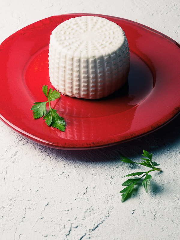 Small ricotta on red dish with parsley on white textured table.
Ricottina fresca su piatto smaltato rosso con prezzemolo su tavolo bianco ruvido