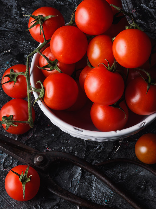 Pomodori ciliegino in un a ciotola bianca su tavolo nero ruvido e forbice da giardino di ferro.
Round tomatoes  in a white bowl on a black textured table and an iron scissor.