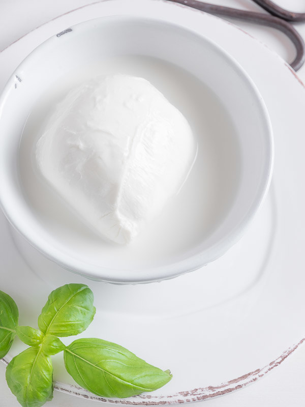 Mozzarella in a white bowl on a white dish with basil leaves.
Mozzarella in uan ciotola bianca su sfondo bianco e foglie di basilico